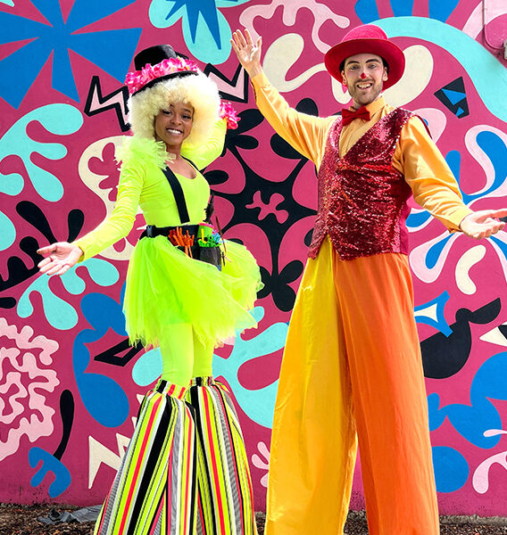 Two vibrant stiltwalkers posing in front of a colorful mural during a live entertainment event in Miami